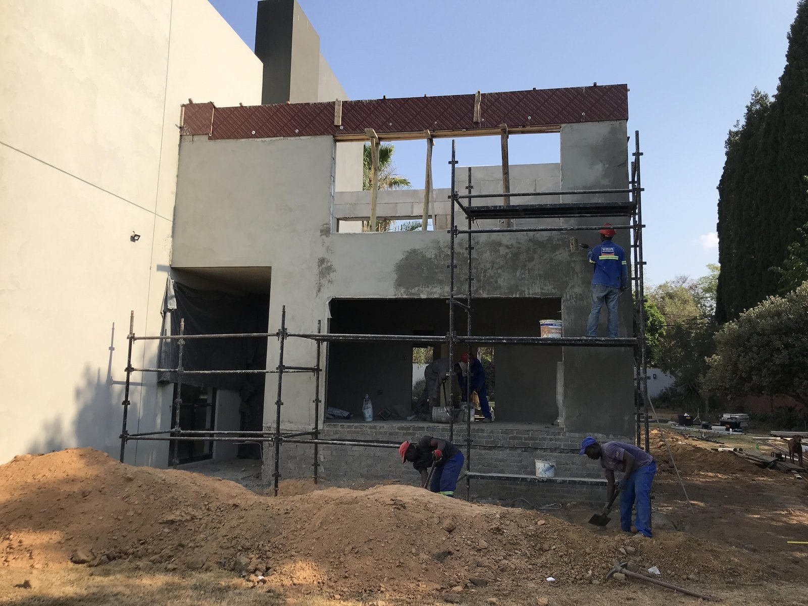 Two-storey house exterior partly rendered, workers plastering from scaffold over block walls.
