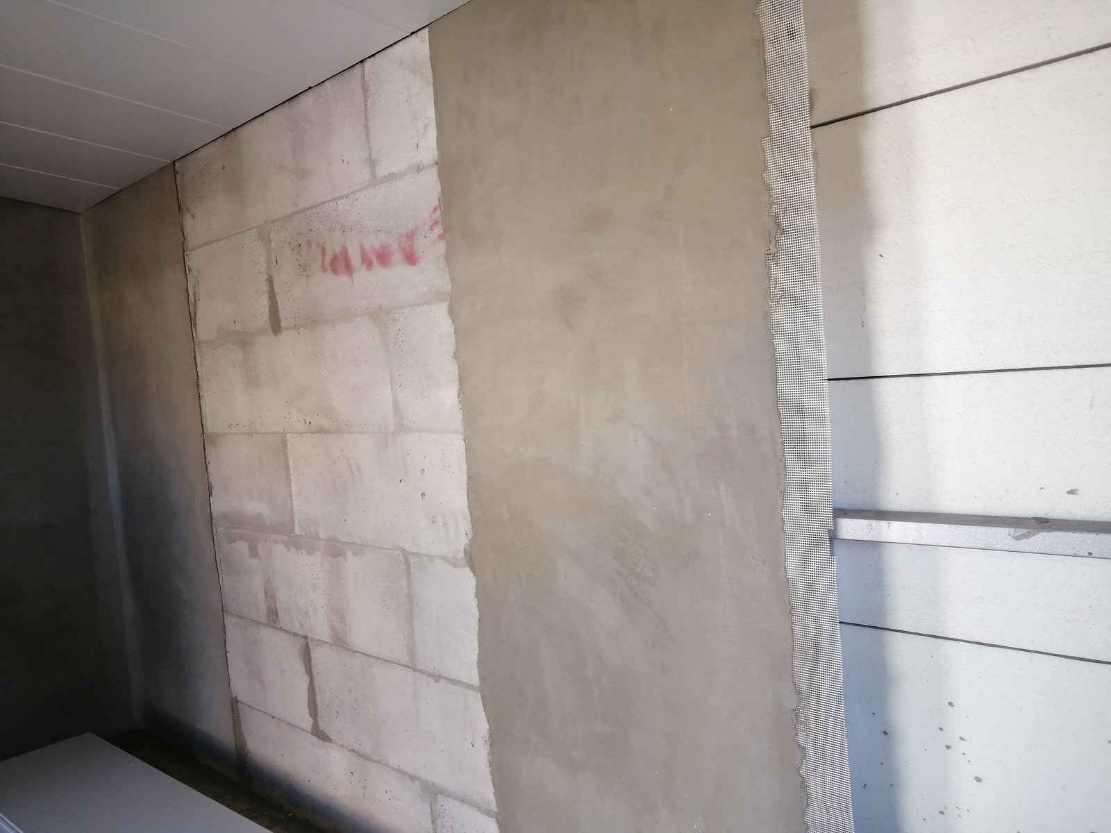 Block wall corner mid-render, reinforcing mesh strip visible next to an adjoining insulated panel.