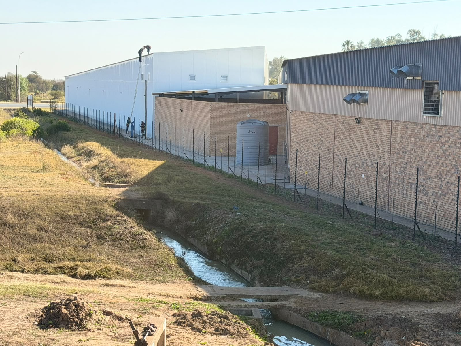 Site view of a long building clad in white micro-ribbed insulated wall panels, with the crew still working off ladders at the top of the elevation. The existing brick and corrugated-steel building stands alongside.