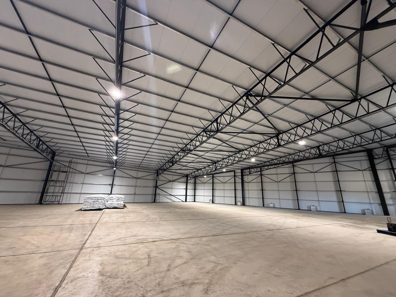 Inside a completed shed: off-white panels line both the walls and the roof soffit, spanning between the black steel trusses and portal frames. Floor is still bare, with a scaffold tower left in place.