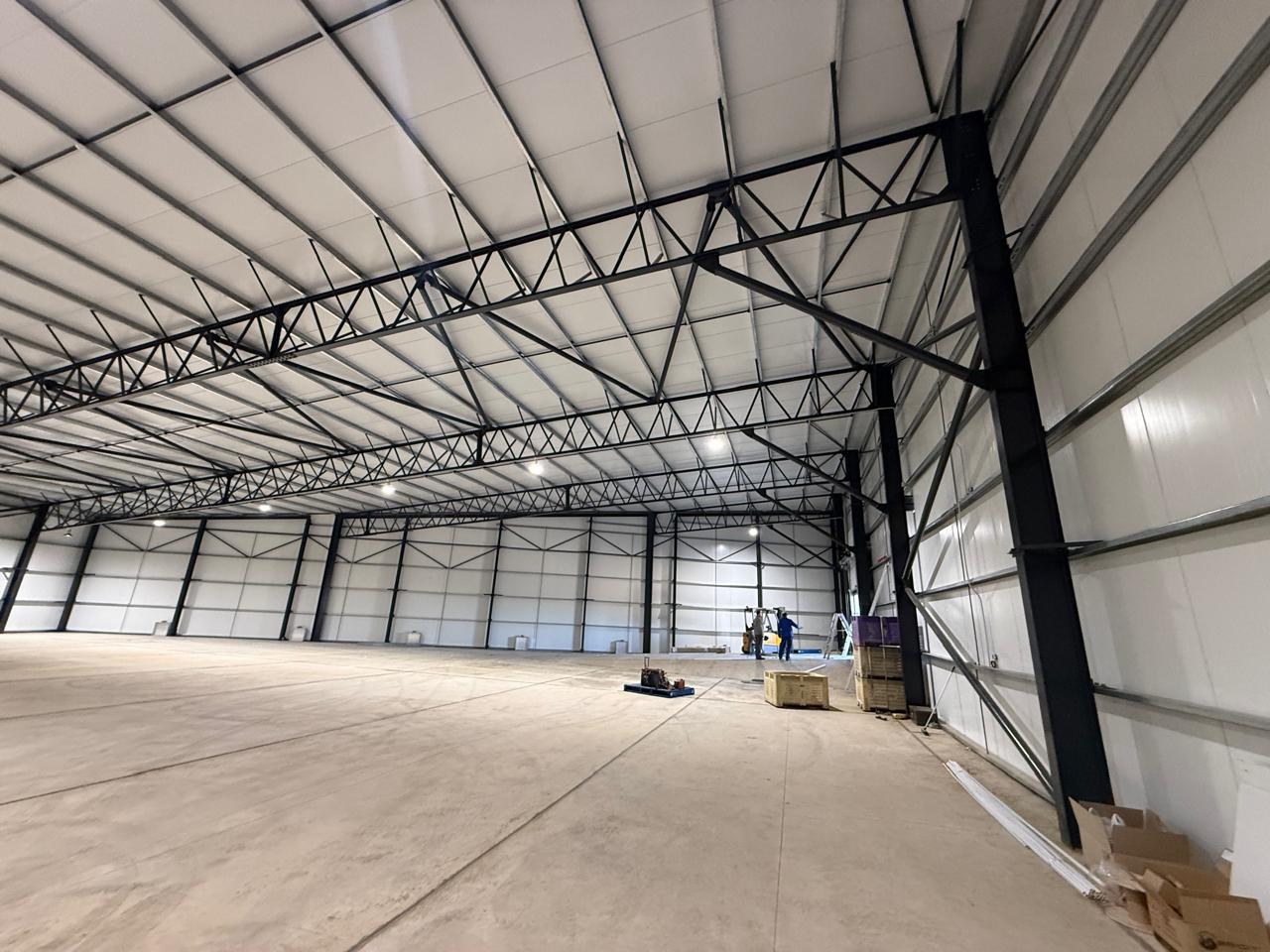 The same building from floor level, with the off-white panel lining running unbroken across the walls and roof soffit behind the steel frame. Two workers are still finishing off in the far bay.