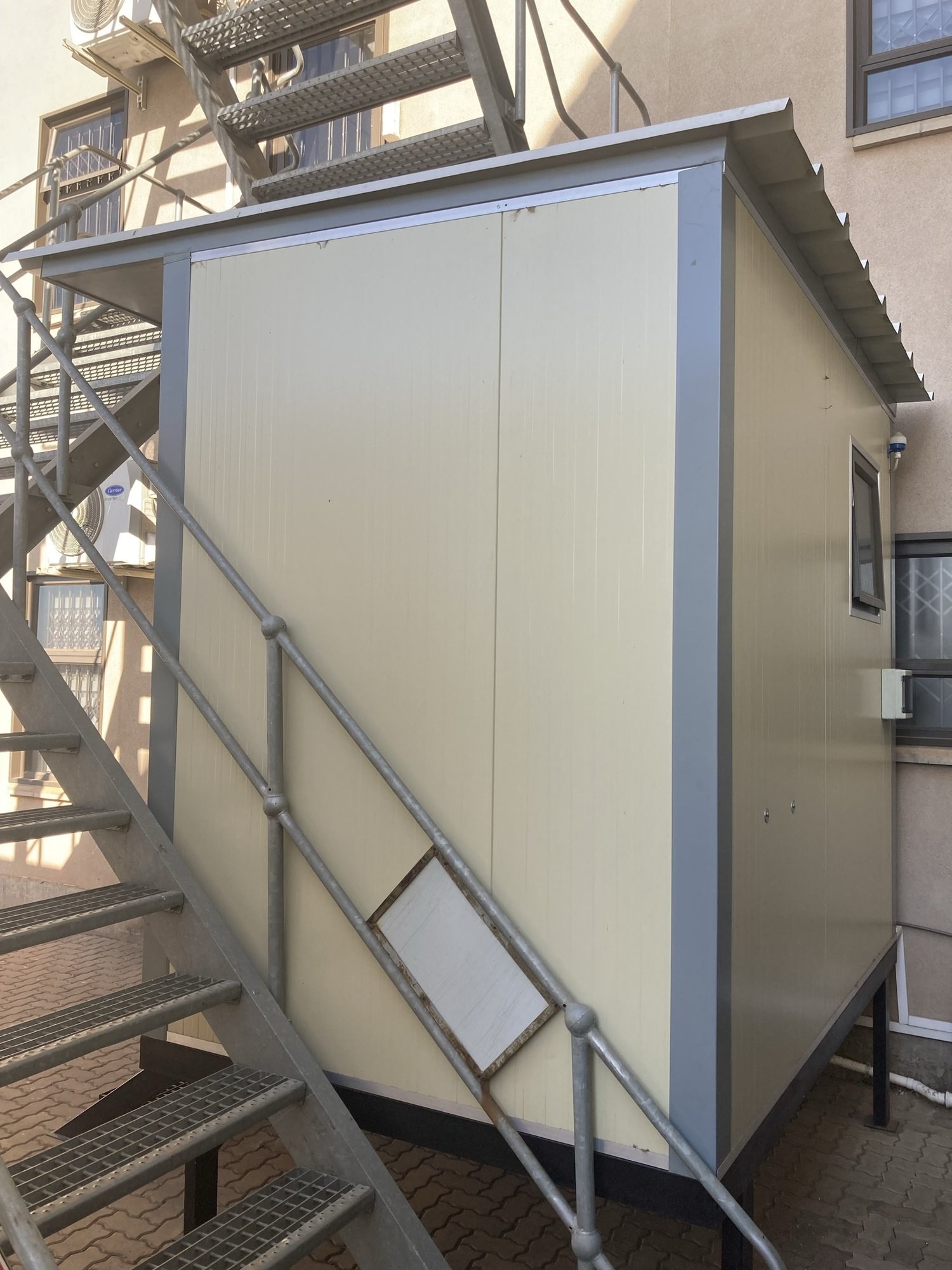 Small demountable cabin built from insulated wall panels, mounted on a raised platform beside a building's exterior stairs