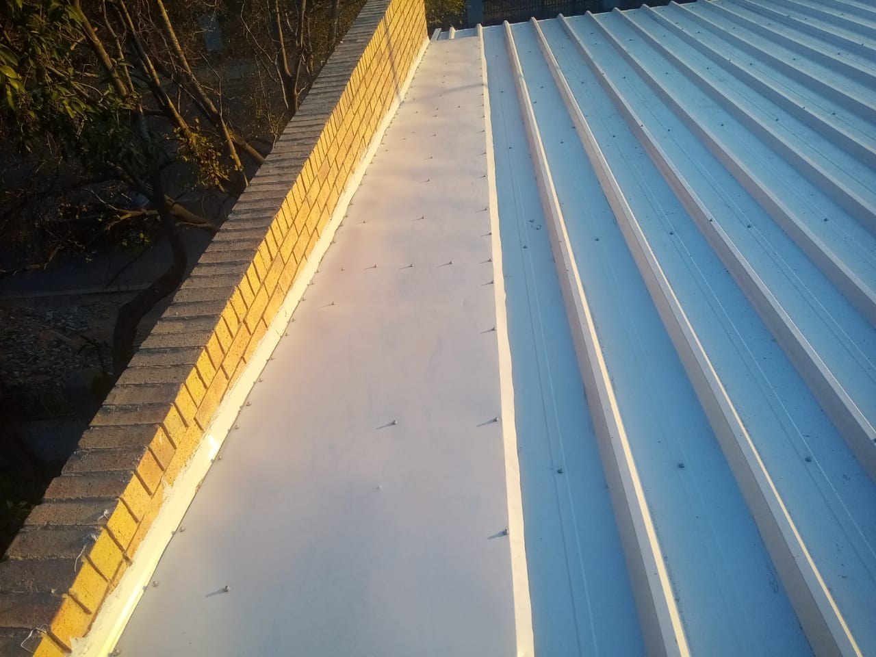 Installed roof at the parapet: ribbed steel sheeting fixed with screws through the flat pans, and a wide flashing turned up against the face-brick parapet wall.