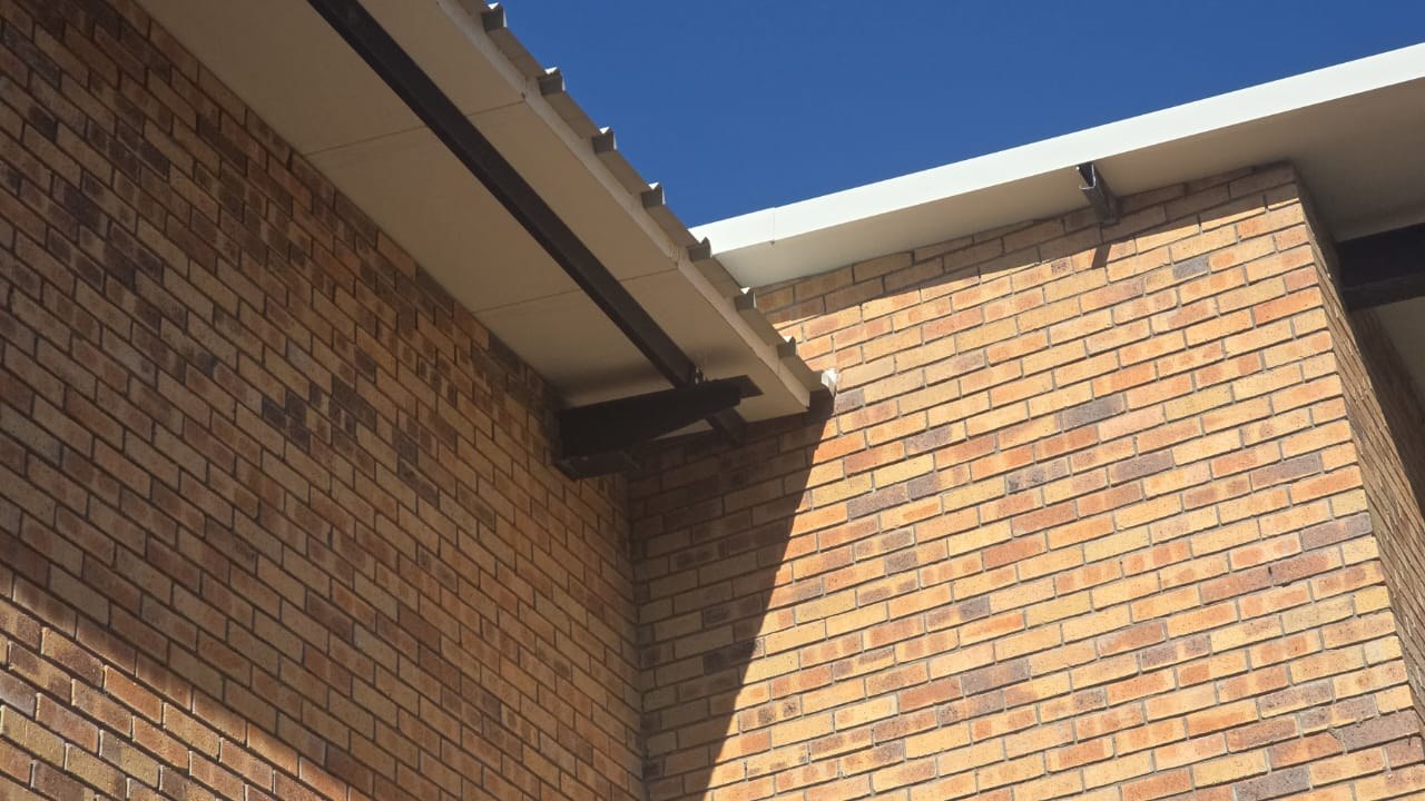 Roof edge over face brickwork: the eave shows the ribs of the steel top sheet in profile with a flat cream underside beneath, carried on a dark steel beam. Most of the frame is the brick wall.