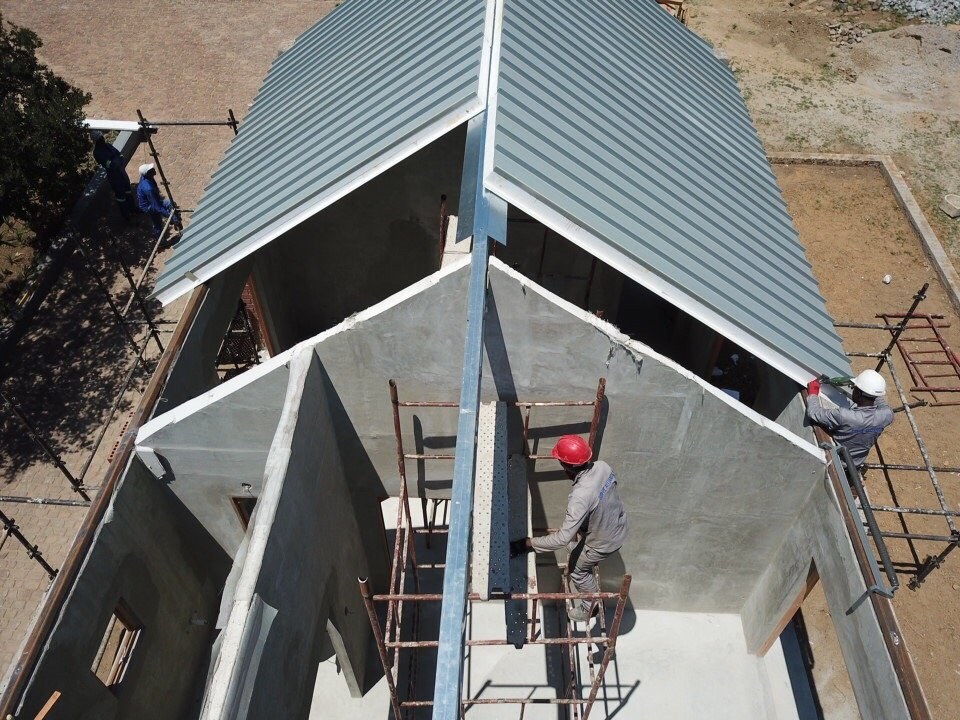 Aerial view of roofers fitting standing-seam sheeting at a hip roof apex, working off scaffolding