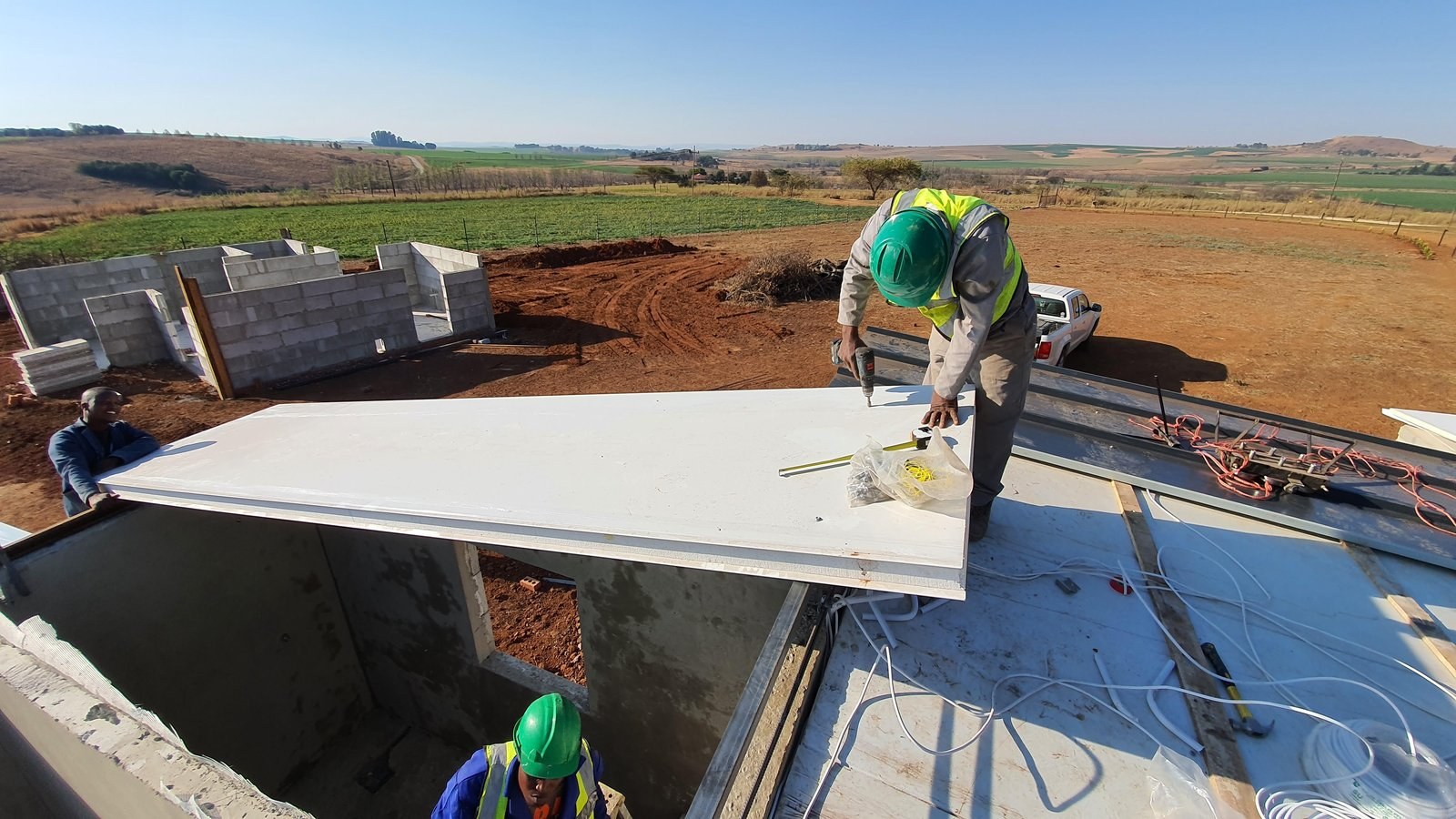 Roofer drilling an insulated roof panel into place on a farmhouse build, open farmland in the background