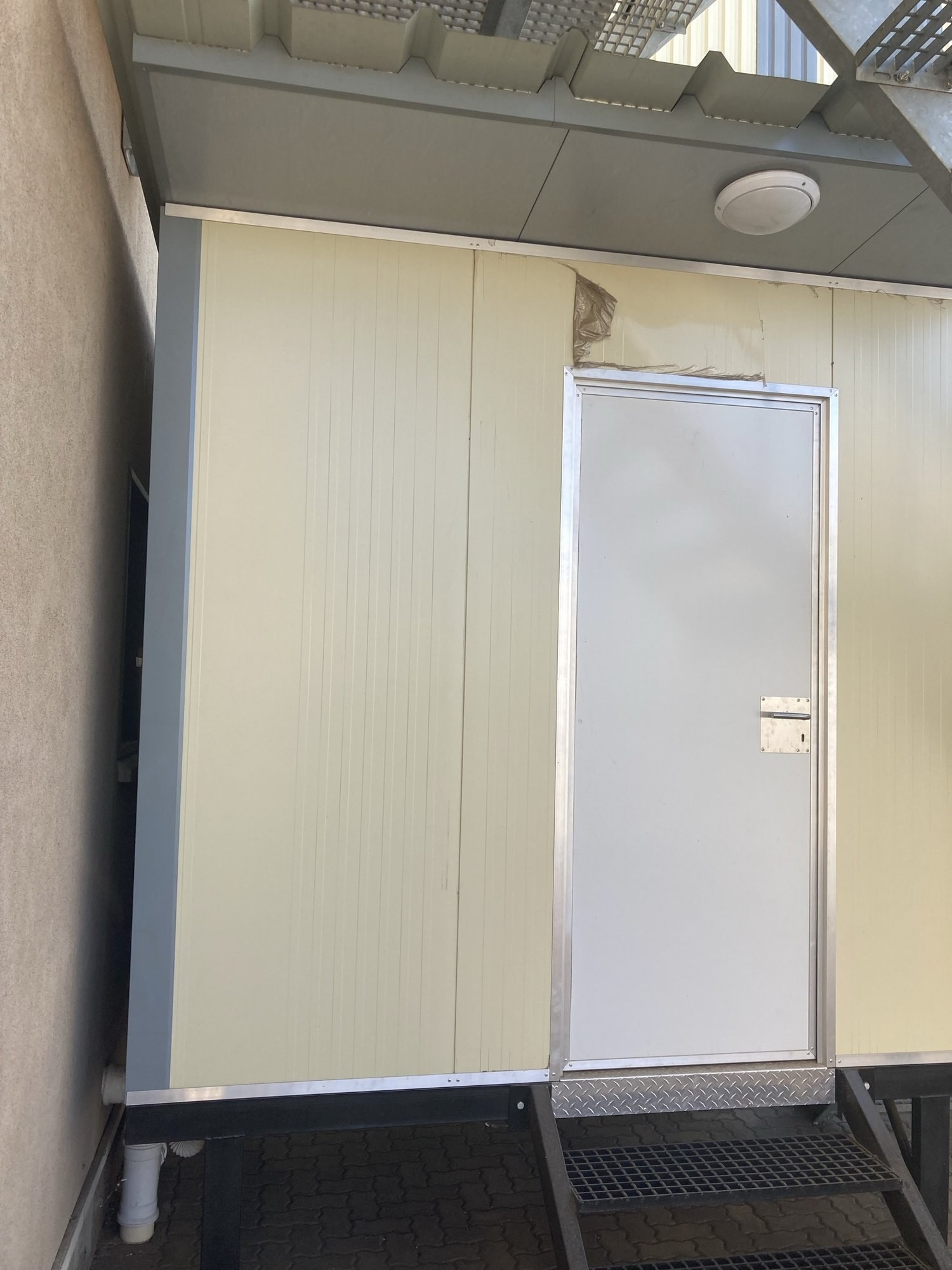 A single white door leaf in an aluminium frame, hung in a cream ribbed sandwich-panel wall under a steel canopy. The panel face above the door head is torn open where it was cut in.