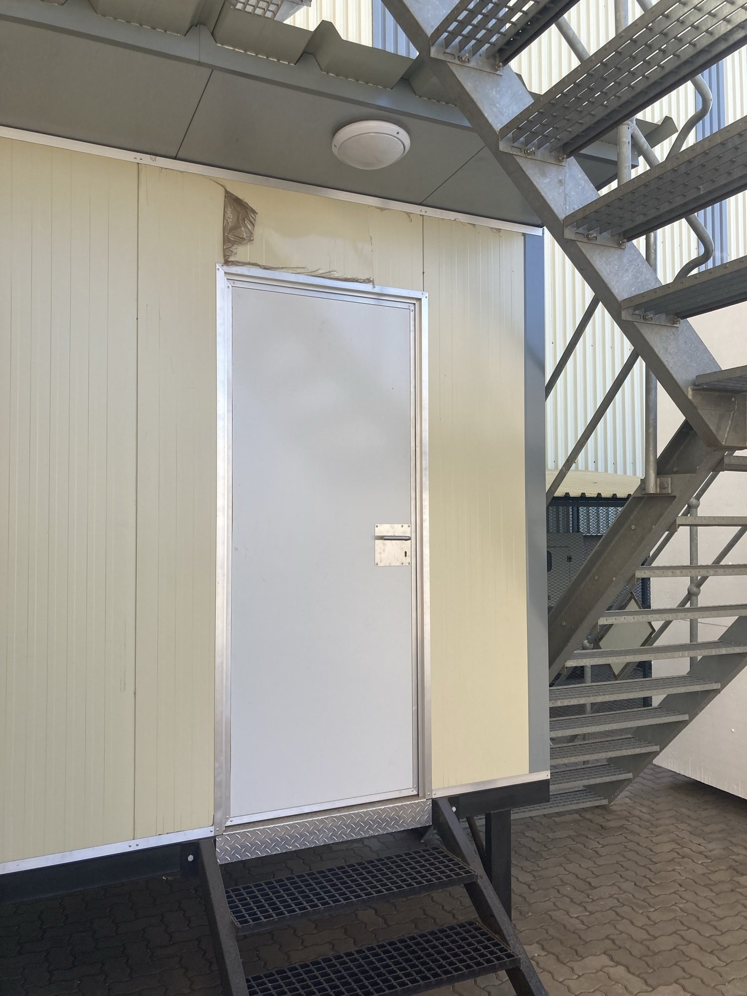 The same white door leaf and aluminium frame in the panel wall, seen from the galvanised access staircase. Grating step and chequer-plate threshold at the sill.