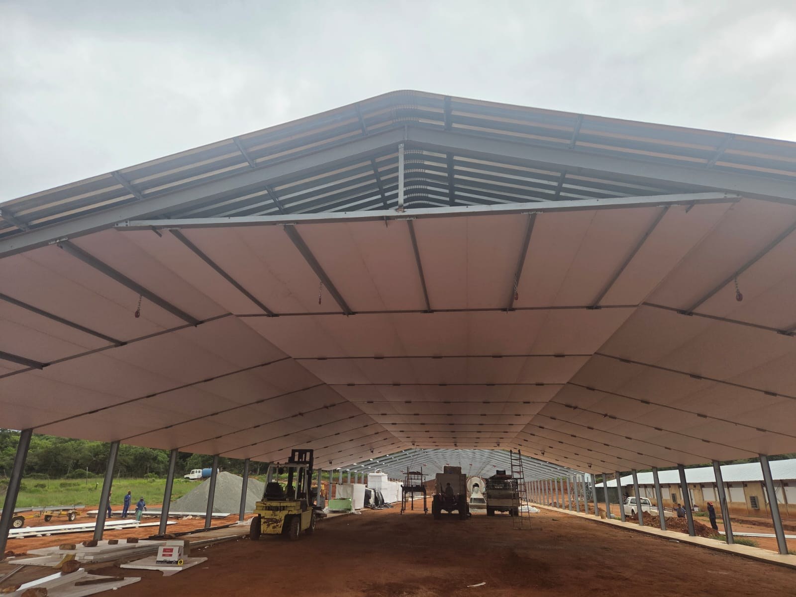 Cream faced panels being installed across both roof slopes of a wide-span agricultural building, with the apex bay still open and stacked boards and a forklift on the ground below.