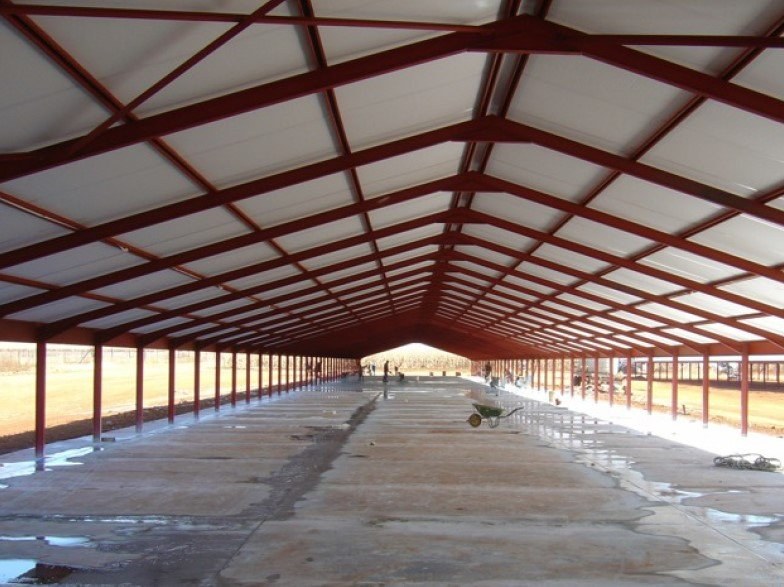 Shed under construction with red steel trusses and insulated ceiling panels overhead, workers moving a wheelbarrow across the wet concrete floor