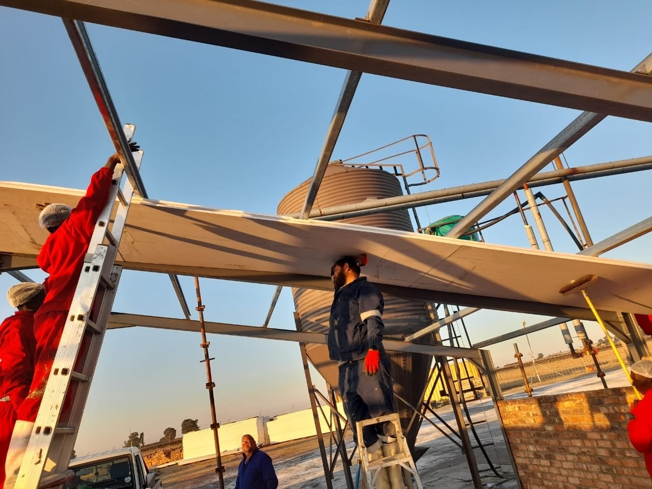 Team installing a long insulated roof panel onto a steel frame beside a feed silo
