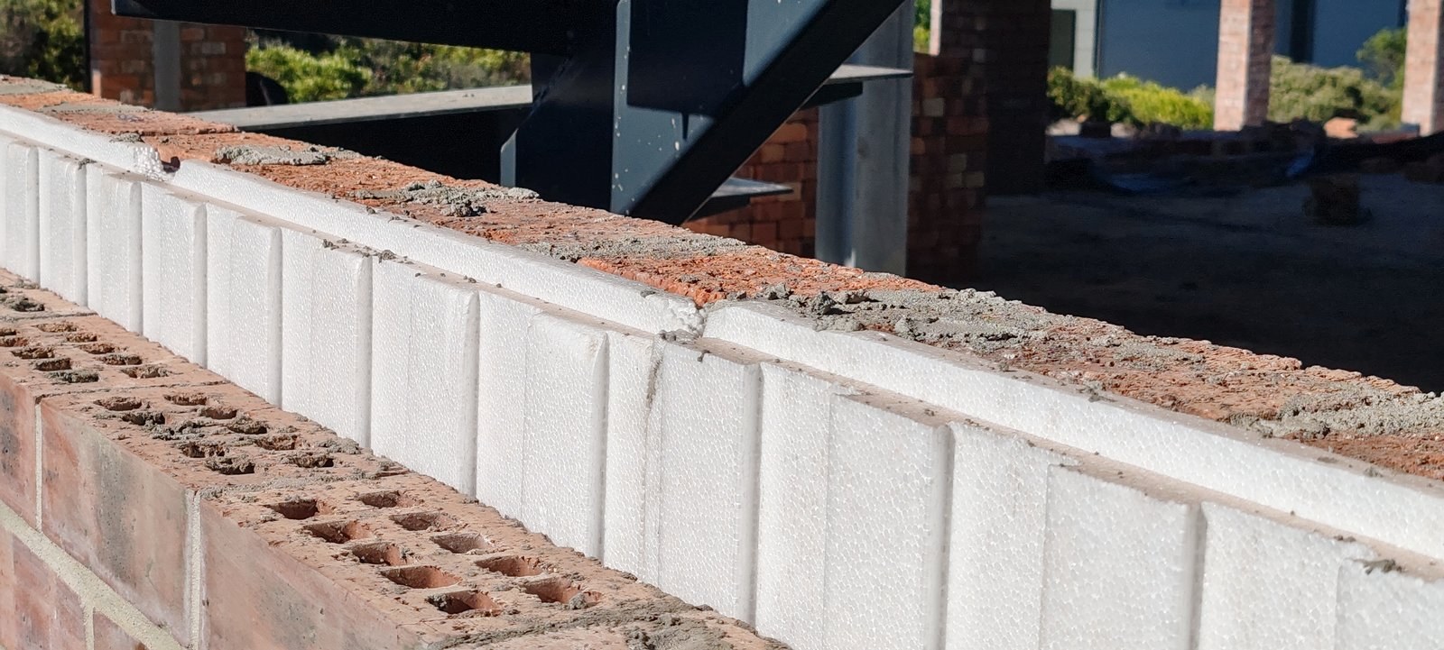 White EPS boards standing on edge in the cavity of a part-built wall, with the perforated clay-brick inner leaf on one side and the mortared brick outer leaf on the other. Steel frame and brick piers of the house behind.