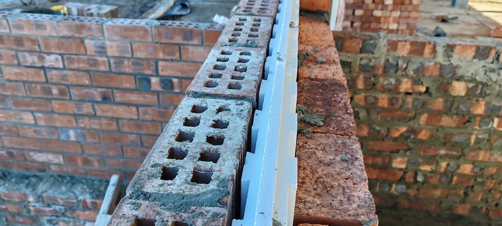 Looking down the top of a cavity wall: the white keyed EPS board sits between a perforated clay-brick leaf and the brick outer leaf, its stepped castellated edge visible along the top.