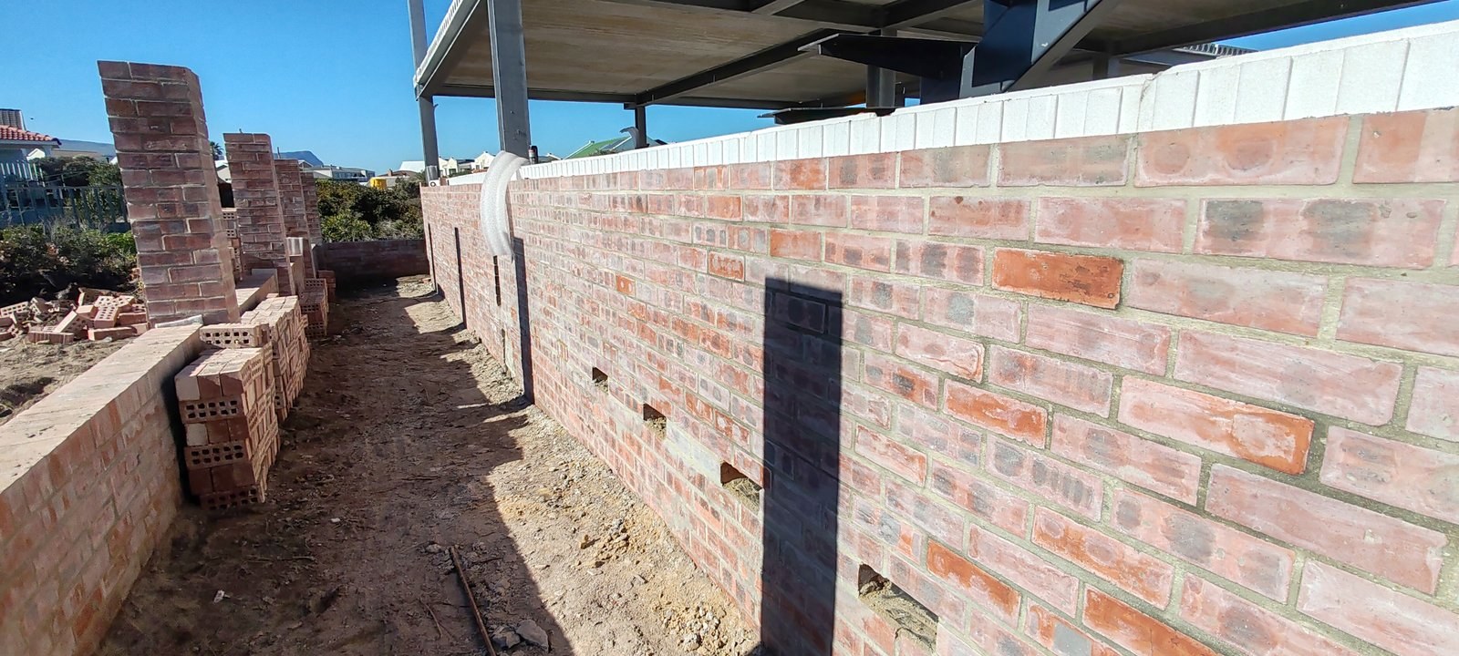 A long run of face brickwork on site with the white EPS boards continuing along the top of the cavity; stacked perforated clay bricks and the steel frame of the house alongside.