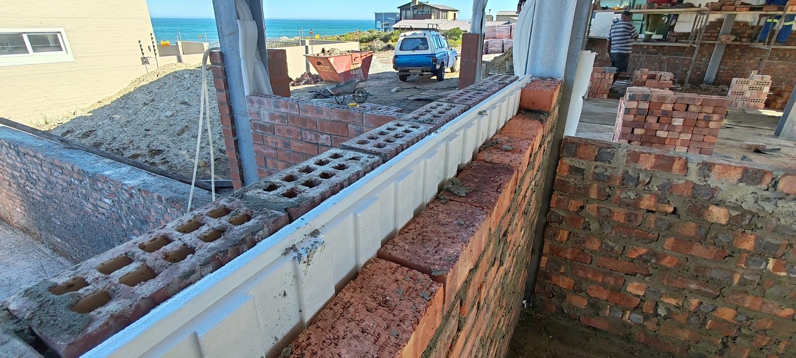 Brick and cavity board boundary wall under construction, coastal site with delivery truck and skip bin