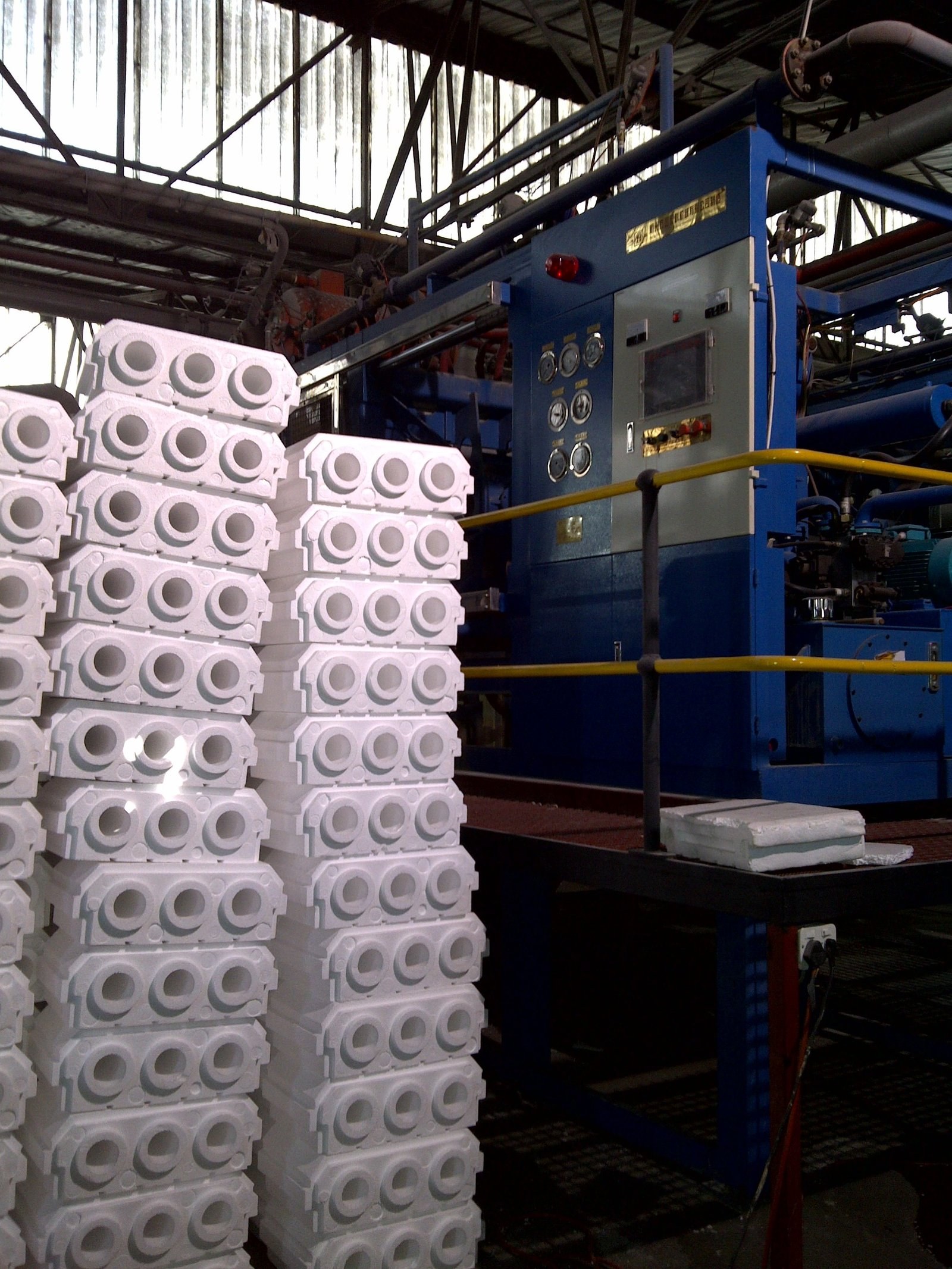 Moulded EPS blocks stacked on the factory floor next to the moulding machine, each block showing its three cores and notched profile.