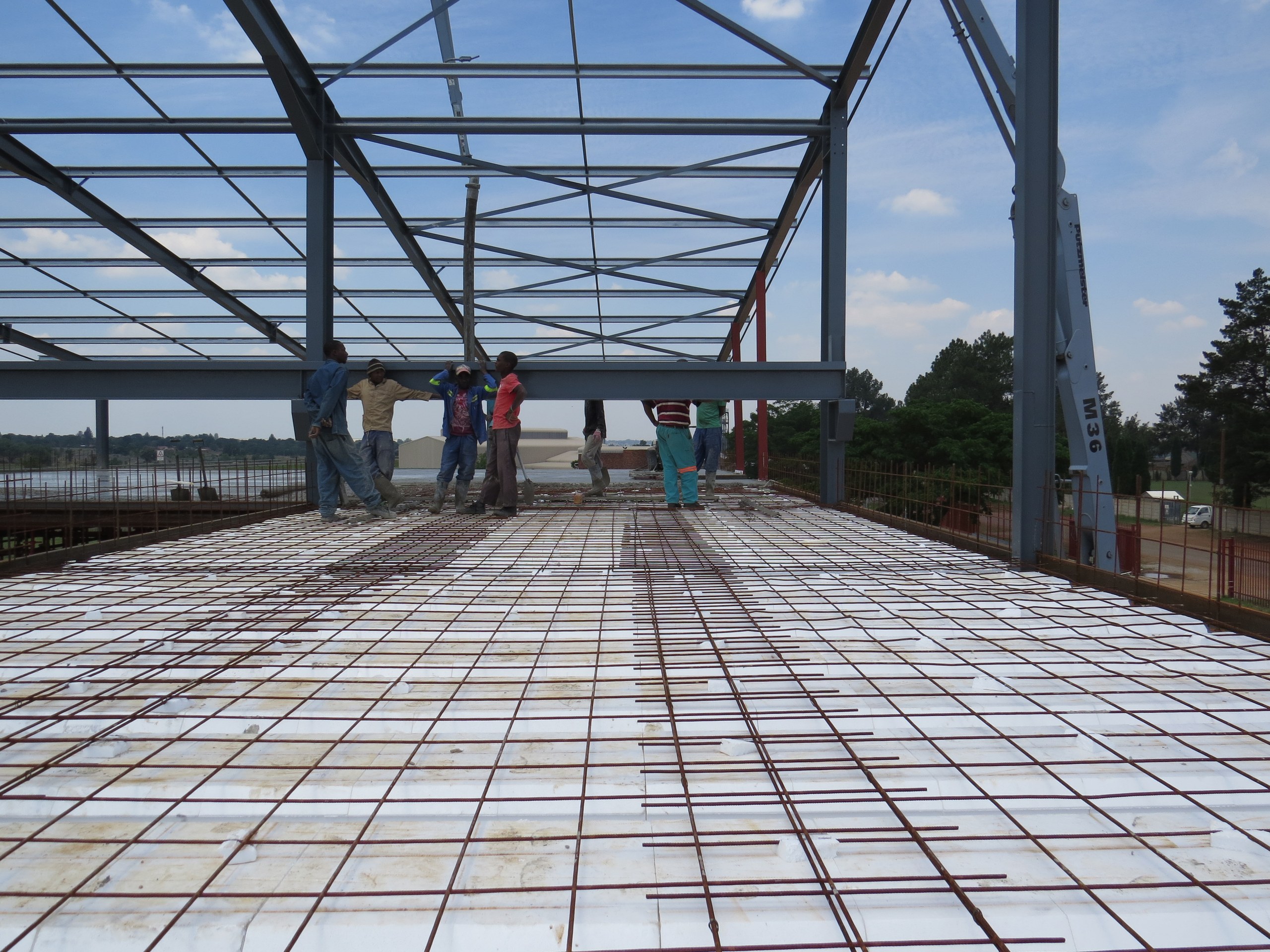 Void formers laid out across the steel frame of a building, with reinforcing bar being placed over them ahead of the pour. The blocks tile the whole floor area; the concrete fills only the ribs between them and a thin topping over the top.