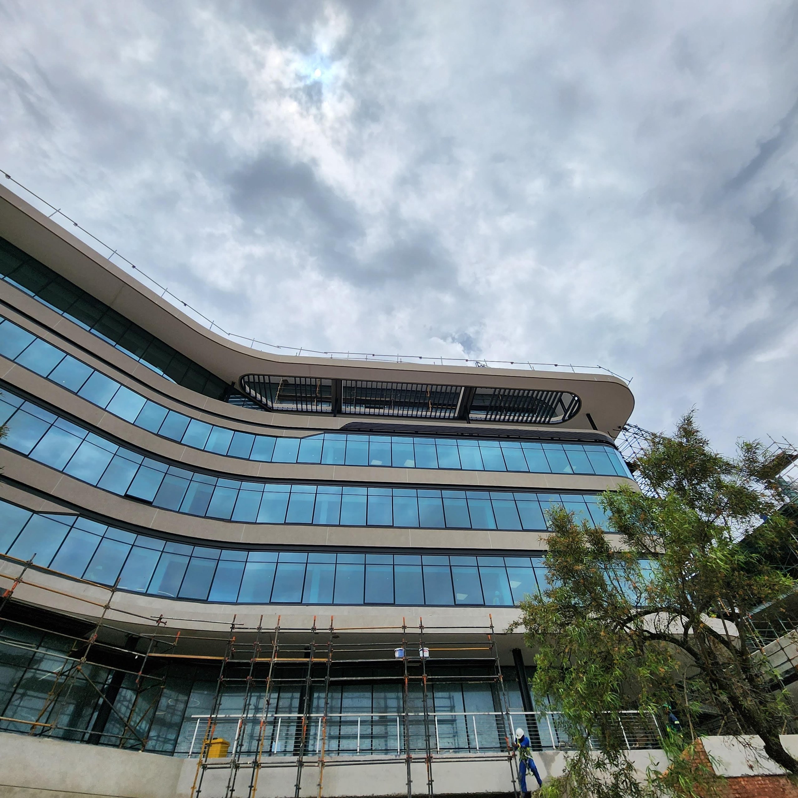 Curved multi-storey facade with smooth pale rendered bands running between the blue ribbon glazing. Scaffolding is still standing at ground level and a worker is on the deck - the coating on the upper floors is complete, the building is not.