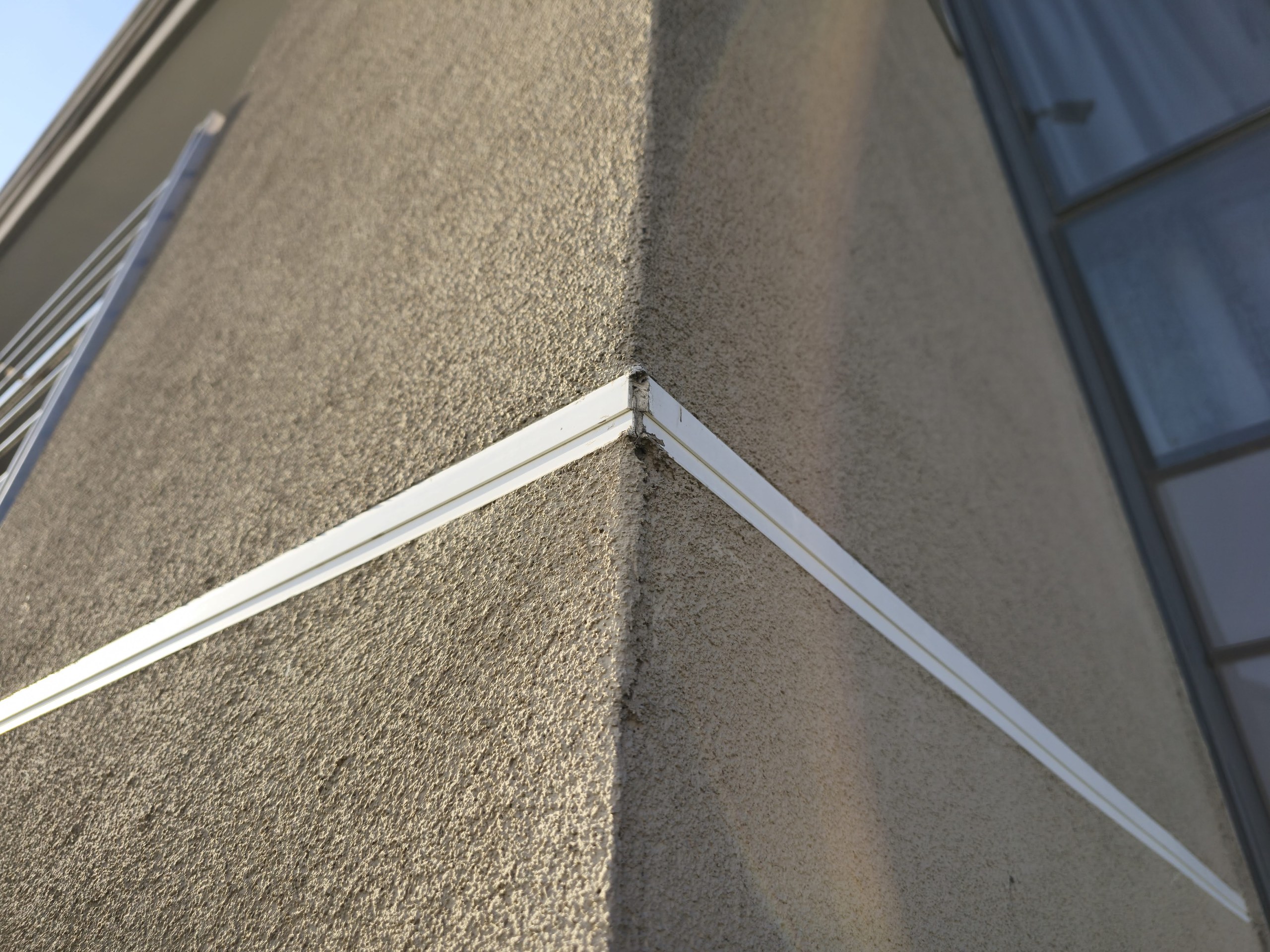 Coarse textured topcoat on an external corner, with a white trim profile turning the arris. Raking low sun shows the aggregate grain of the finish; the render on the corner edge itself is slightly chipped.