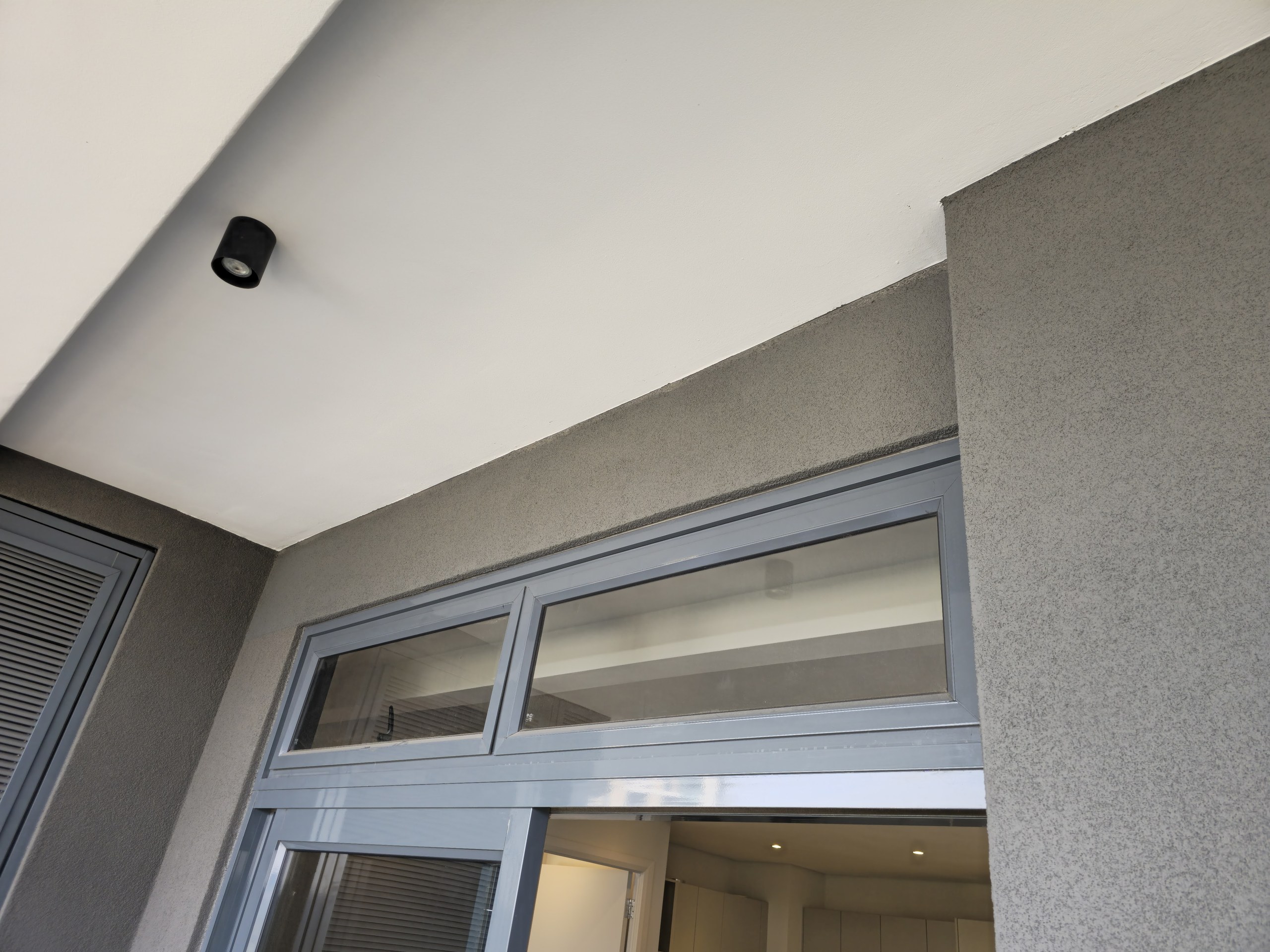 Underside of a balcony: smooth white soffit meeting grey textured render on the wall and pier, with the coating run cleanly down to the aluminium sliding-door head. Shot from outside, looking in through the open door.