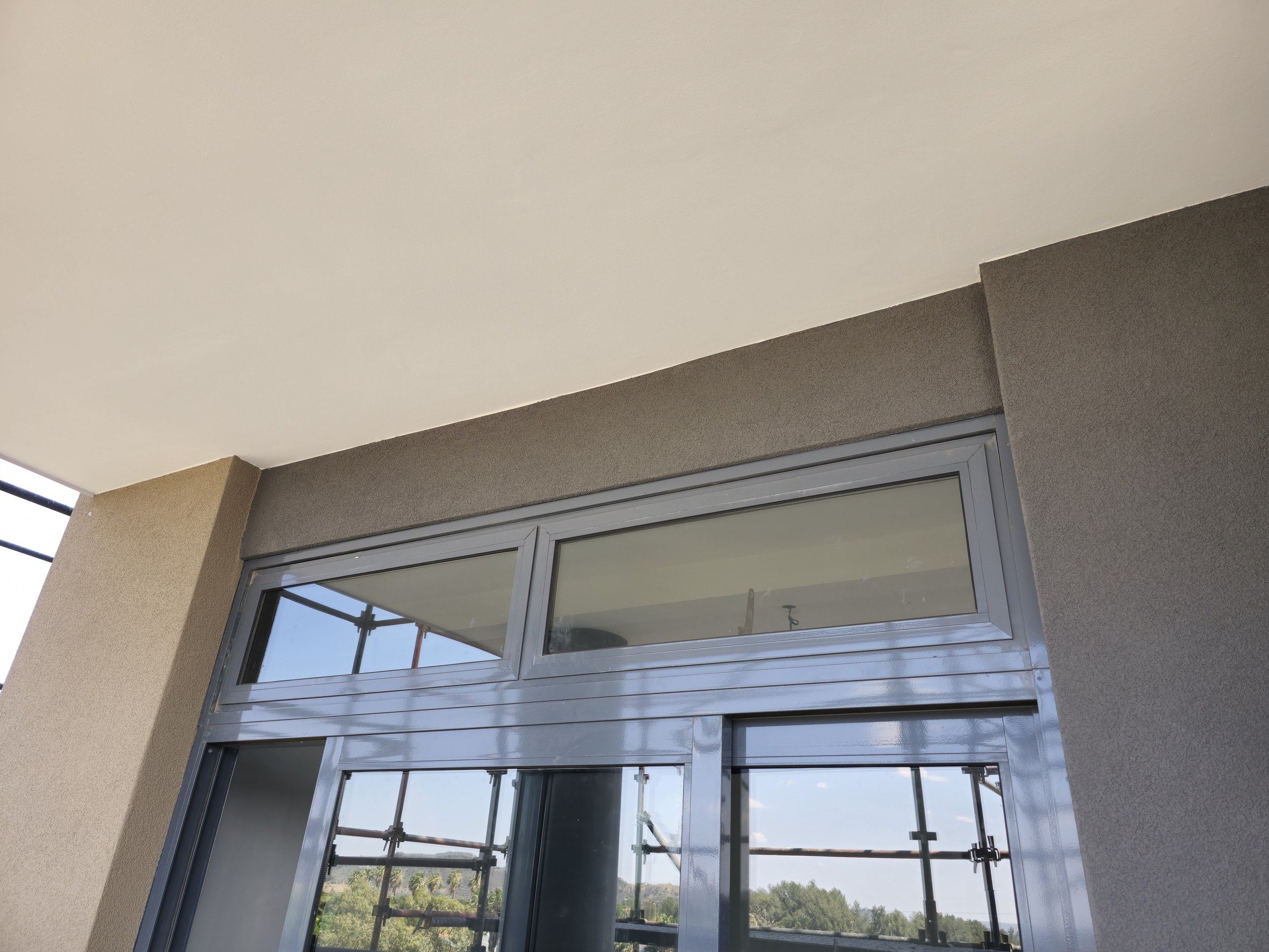 Textured render on the beam face and pier meeting a smooth painted soffit above an aluminium sliding door. Scaffolding is still standing outside, visible through the glass.
