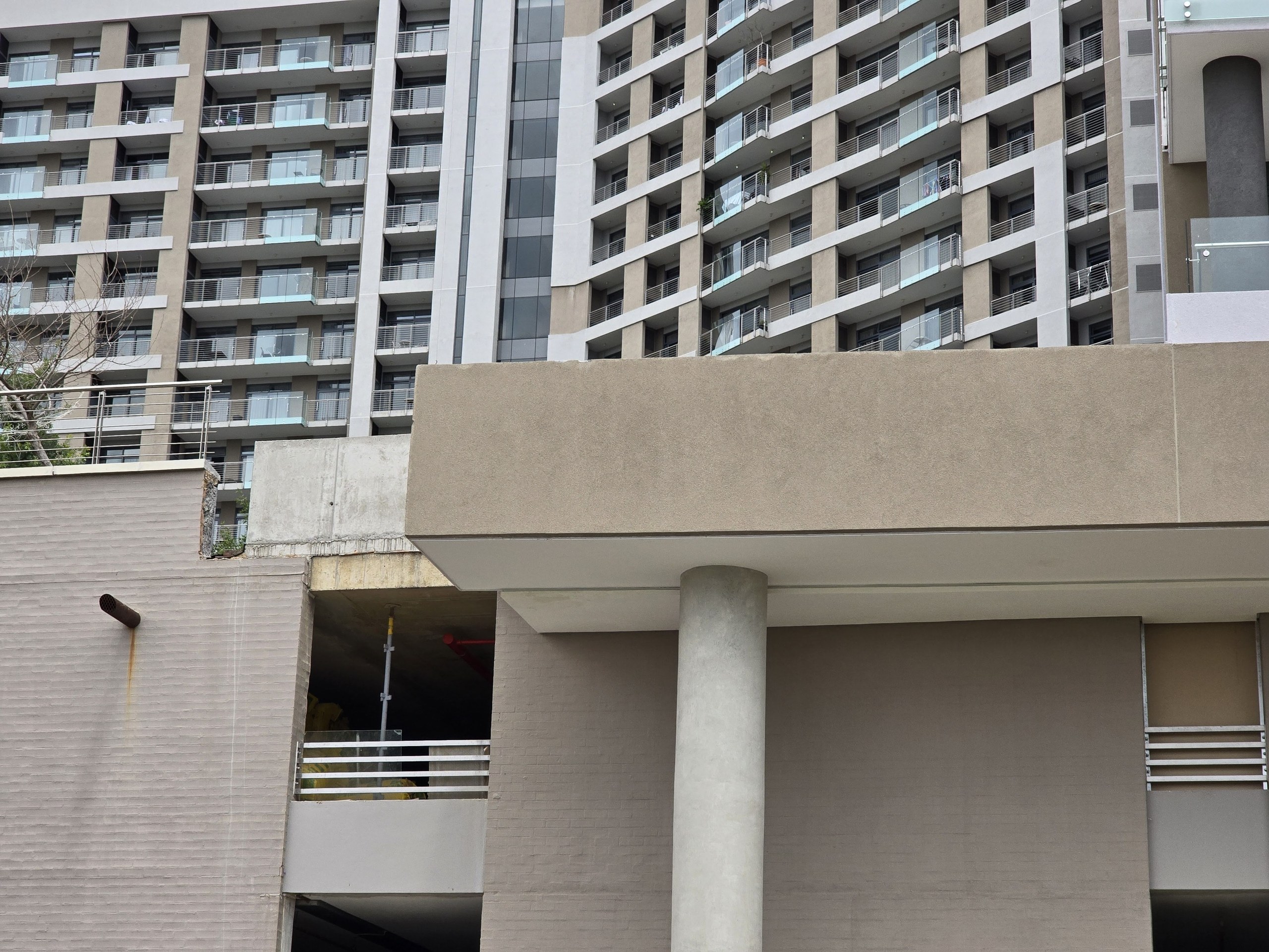 Rendered podium under a multi-storey apartment block, photographed mid-construction. A round rendered column carries a coated soffit and a thick band of beige textured render; the wall to the left is finished in a horizontally grooved texture. The site is still working: bare concrete at the slab edge, an open parking undercroft with a prop and a worker in yellow, and a rust stain running down from a steel pipe outlet.