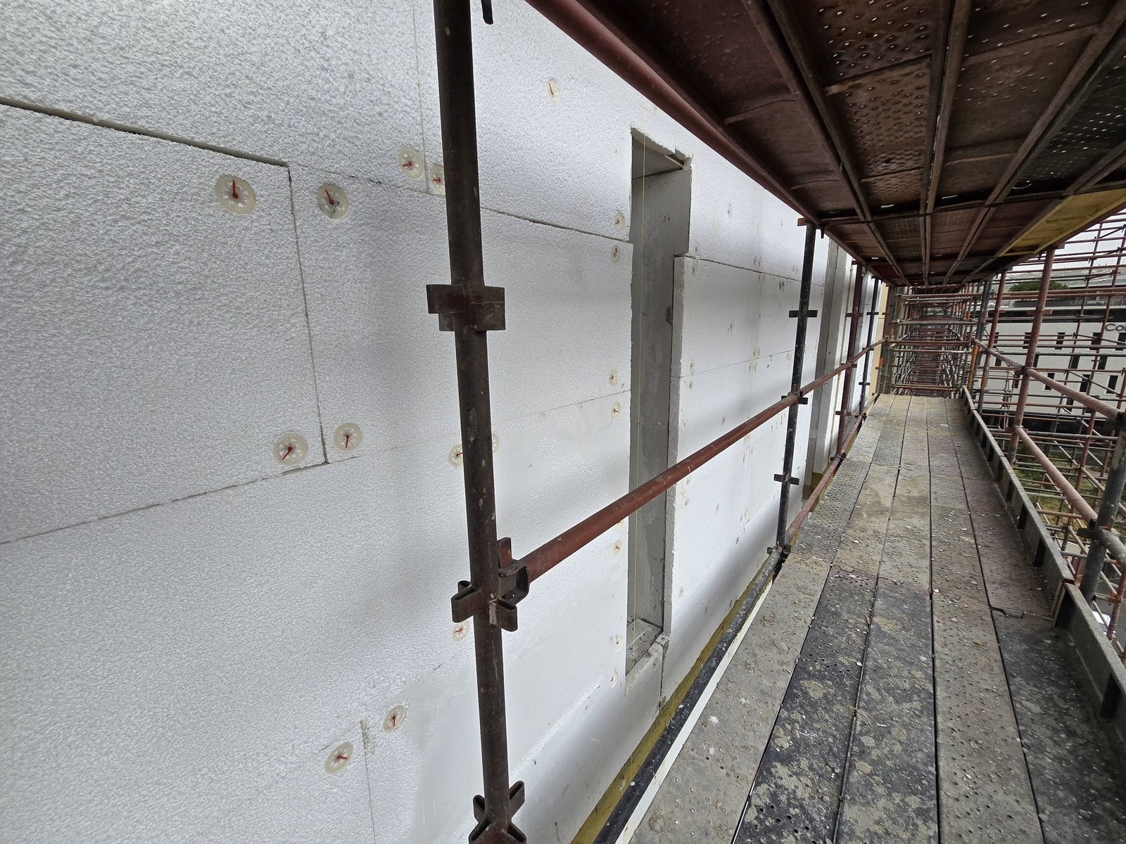 EPS boards fixed to a high-rise facade in a running-bond pattern with disc fixings, scaffold walkway alongside
