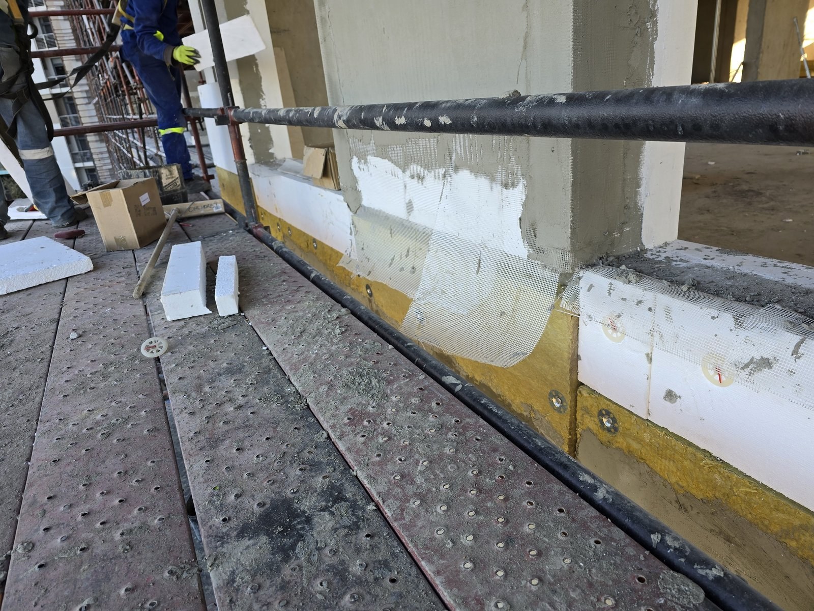 Mesh and basecoat applied over EPS boards at a column corner, foam offcuts left on the scaffold deck